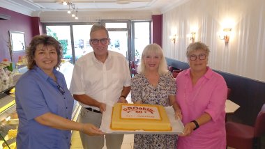 Vier Personen lächeln und halten gemeinsam einen Kuchen mit der Aufschrift "PROaktiv 60+ Zum 1. Geburtstag". Sie stehen in einem gemütlichen Café mit rot-weißen Wänden und gedeckten Tischen.