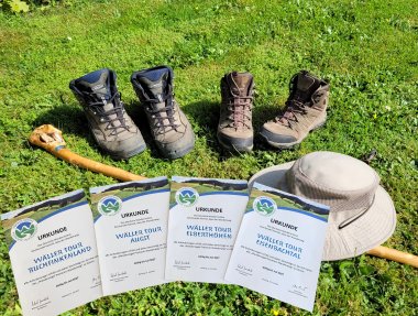 Das Bild zeigt vier Zertifikate für erfolgreiche Wanderungen ("Wäller Tour Buchfinkenland", "Wäller Tour Augst", "Wäller Tour Elberthöhen", "Wäller Tour Eisenbachtal") auf grünem Rasen, zusammen mit zwei Paar Wanderschuhen, einem Wanderstock und einem beigefarbenen Sonnenhut. Die Szene symbolisiert die Erfolge und Ausrüstungen eines Wanderers.