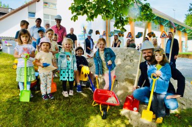 Eine Gruppe von Kindern und Erwachsenen posiert fröhlich beim Spatenstich vor der Kita Pusteblume im Freien. Die Kinder tragen bunte Kleidung und halten Schaufeln und eine Schubkarre, während einige Erwachsene im Hintergrund Bauhelme tragen. Im Hintergrund ist die Kita mit Baustellenschildern und Spielzeug-Baumaschinen zu sehen.