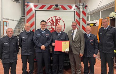 Eine Gruppe von sieben Männern posiert in einer Feuerwehrwache vor einem Feuerwehrfahrzeug mit rot-weißen Warnstreifen und einem Feuerwehr-Logo auf dem Rollladen. Die meisten Männer tragen dunkelblaue Feuerwehruniformen mit Namensschildern und Abzeichen, einer von ihnen hält eine Urkunde in einer roten Mappe. Ein Mann in der Mitte trägt einen Anzug mit Krawatte. Alle lächeln in die Kamera, die Szene deutet auf eine Ehrung oder Ernennung innerhalb der Feuerwehr hin.