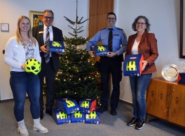 Ein Gruppenfoto mit vier Personen vor einem geschmückten Weihnachtsbaum. Alle halten leuchtend gelbe Fahrradhelme, teilweise in Verpackungen, während weitere Helme am Boden gestapelt sind. Die Szene scheint eine Spenden- oder Geschenkübergabe zu sein, möglicherweise für eine Verkehrsschule oder eine ähnliche Initiative.