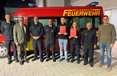 Eine Gruppe von neun Personen steht vor einem roten Feuerwehrfahrzeug mit der Aufschrift "HEILIGENROTH FEUERWEHR". Zwei junge Menschen in der Mitte halten rote Urkunden in der Hand. Die Gruppe besteht aus Feuerwehrmitgliedern in Uniform, einer Person im Anzug und einer Person in Freizeitkleidung.