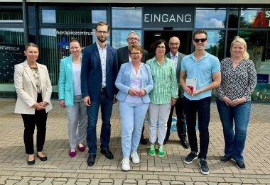 Eine Gruppe von neun Personen steht vor dem Eingang eines Therapiezentrums, darunter vier Frauen und fünf Männer. Die Gruppe trägt formelle bis halb-formelle Kleidung und posiert lächelnd für das Foto. Im Hintergrund sind die Schriftzüge "Therapiezentrum" und "Eingang" zu sehen.