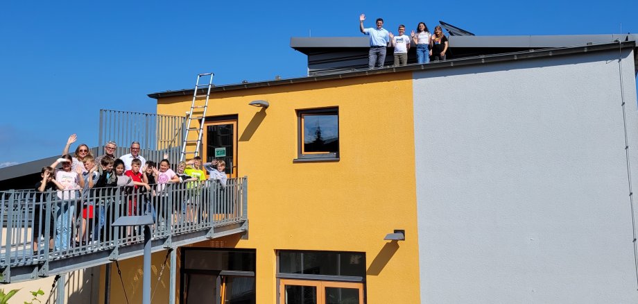 Eine Gruppe von Kindern und Erwachsenen steht auf einem Balkon und einer Dachterrasse eines gelb-grauen Gebäudes und winkt in die Kamera. Ein Leiter führt vom Balkon zum Dach, wo weitere Personen stehen und ebenfalls winken. Der Himmel ist klar und blau.