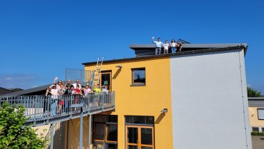 Eine Gruppe von Kindern und Erwachsenen steht auf einem Balkon und einer Dachterrasse eines gelb-grauen Gebäudes und winkt in die Kamera. Ein Leiter führt vom Balkon zum Dach, wo weitere Personen stehen und ebenfalls winken. Der Himmel ist klar und blau.