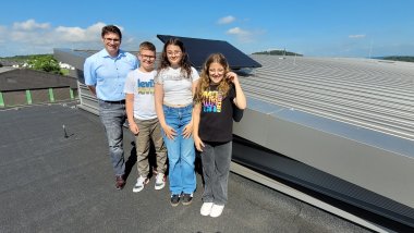 Ein Mann und drei Kinder stehen auf einem Dach neben einer Solaranlage und lächeln in die Kamera. Der Himmel ist blau mit einigen Wolken, und im Hintergrund sind andere Gebäude und Bäume zu sehen. Die Kinder tragen lässige Kleidung und der Mann trägt ein hellblaues Hemd und Jeans.