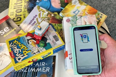Eine Hand hält ein Smartphone, auf dem die PayPal-App geladen wird. Im Hintergrund liegen mehrere Bücher und ein kleines Spielzeugauto auf einem Tisch verstreut.