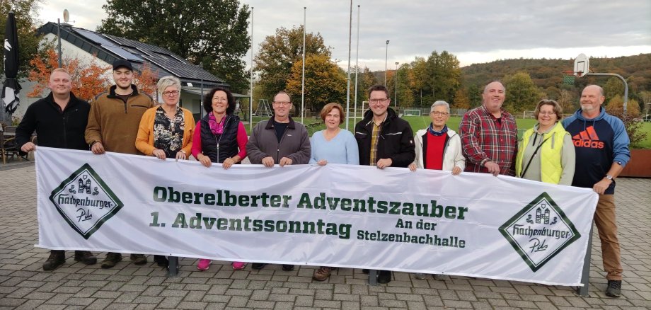 Eine Gruppe von elf Personen steht draußen und hält ein Banner mit der Aufschrift: „Oberelberter Adventszauber, 1. Adventssonntag an der Stelzenbachhalle“. Im Hintergrund sind ein Gebäude mit Solarpanels, herbstliche Bäume und ein Sportplatz zu sehen. Das Banner trägt die Logos von „Hachenburger Pils“.