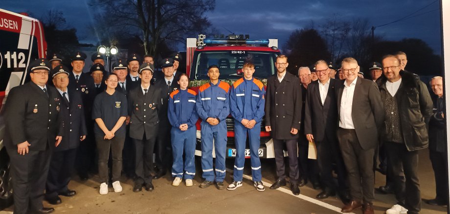 Eine Gruppe von Feuerwehrleuten und jungen Feuerwehranwärtern steht in Uniform vor einem Feuerwehrfahrzeug, begleitet von mehreren Personen in Zivilkleidung. Die Aufnahme scheint am Abend gemacht worden zu sein, vor einem dunklen Himmel, mit beleuchteten Feuerwehrfahrzeugen im Hintergrund. Alle blicken lächelnd in die Kamera, was auf eine feierliche Veranstaltung, möglicherweise die Übergabe eines neuen Fahrzeugs, hinweist.