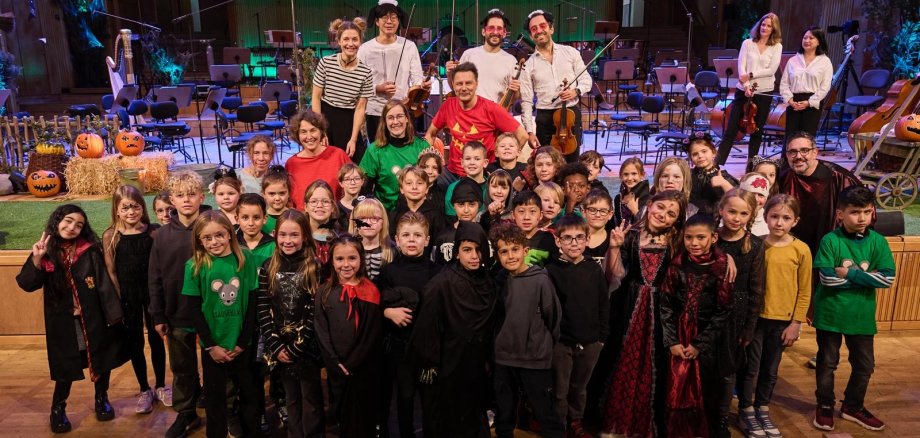 Eine Gruppe von Kindern in Halloween-Kostümen posiert mit Erwachsenen in einem Konzertsaal vor einer Bühne. Im Hintergrund sind ein Orchesteraufbau, Kürbisse, Strohballen und eine herbstlich dekorierte Kulisse zu sehen. Die Stimmung wirkt fröhlich und festlich, passend zum Anlass.