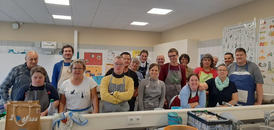 Das Bild zeigt eine Gruppe von Teilnehmenden eines inklusiven Tandem-Kochkurses, bei dem Menschen mit und ohne Behinderung gemeinsam kochen. Die fröhliche Atmosphäre im Kursraum deutet auf das gemeinsame Lernen und das Miteinander hin. Alle Teilnehmenden tragen Schürzen und stehen in einer Küche.