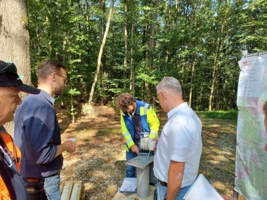 Mehrere Männer stehen im Wald um ein Bohrgerät herum, einer von ihnen führt eine Messung an dem Gerät durch. Einige der Männer halten Notizbücher und verfolgen den Prozess aufmerksam. Rechts im Bild hängt eine Landkarte an einem Gestell, und die Szene spielt sich unter sonnigen Bedingungen ab.