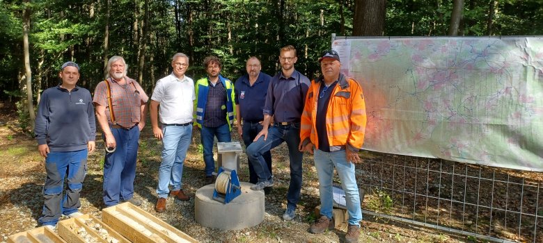 Auf dem Bild stehen sieben Männer in Arbeitskleidung im Wald neben Bohrkern-Proben, die in Holzkisten auf dem Boden liegen. Hinter ihnen hängt eine große Landkarte. Sie scheinen an einem technischen oder geologischen Projekt zu arbeiten, möglicherweise im Zusammenhang mit Erkundungsbohrungen, da auch ein Bohrgerät sichtbar ist. Die Szene ist bei sonnigem Wetter aufgenommen.