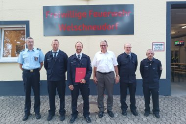 Sechs Männer, darunter fünf Feuerwehrmänner in Uniform, stehen vor dem Gebäude der Freiwilligen Feuerwehr Welschneudorf. Einer der Männer hält eine rote Urkunde. Im Hintergrund ist ein Schild mit der Aufschrift „Freiwillige Feuerwehr Welschneudorf“ zu sehen.