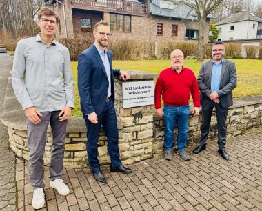 Vier Männer stehen vor einem Gebäude neben einem Schild mit der Aufschrift 'MVZ LandarztPlus-Welschneudorf Facharztpraxis für Allgemeinmedizin.' Die Szene spielt sich auf einem gepflasterten Weg vor einer Steinmauer und einer Rasenfläche ab, mit Wohnhäusern im Hintergrund.