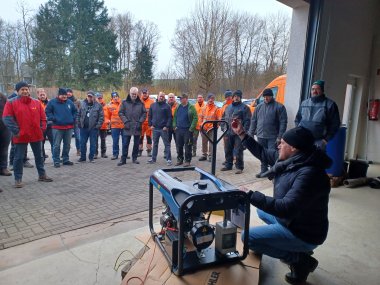 Ein Mann in dunkler Winterkleidung kniet vor einem Notstromaggregat und erklärt es einer Gruppe von Menschen, die in einer offenen Garage stehen. Die Zuschauer, viele in orangefarbener Arbeitskleidung, hören aufmerksam zu. Im Hintergrund sind Bäume, ein oranger Transporter und eine Feuerlöscherhalterung an der Wand zu sehen.