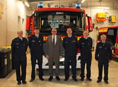  Die Aufnahme zeigt sechs Männer, darunter fünf Feuerwehrmänner in dunkler Uniform und eine Person im Anzug, die in einer Feuerwehrhalle vor einem großen roten Feuerwehrauto mit der Kennung "53/44-1" posieren. Im Hintergrund sind ein weiteres Feuerwehrfahrzeug mit der Aufschrift "Niederelbert" sowie verschiedene Ausrüstungsgegenstände sichtbar.